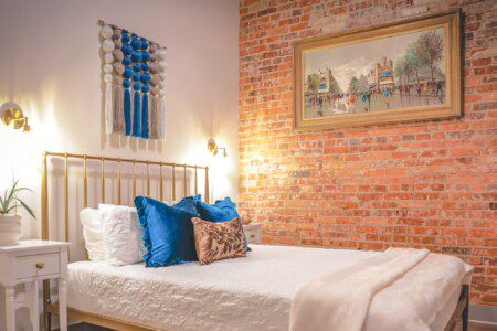Brass bed with blue accent pillows, a macrame wall hanging, and exposed brick