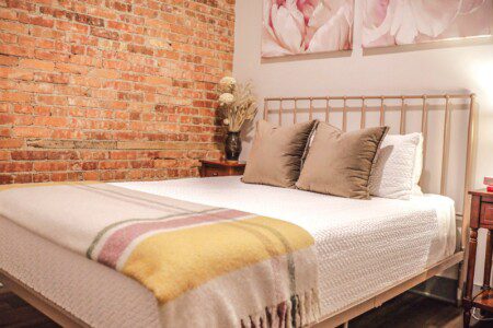 Queen bed dressed in neutral linens with a yellow throw against an exposed brick wall